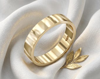 May include: A gold wedding band with a flat design, resting on a white cloth. The ring is marked "14K". A gold leaf sprig is next to the ring. The ring is a symbol of commitment and love.