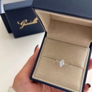 May include: A marquise cut diamond engagement ring with a delicate band and small accent diamonds. The ring is in a blue velvet ring box with the Gioielli logo.