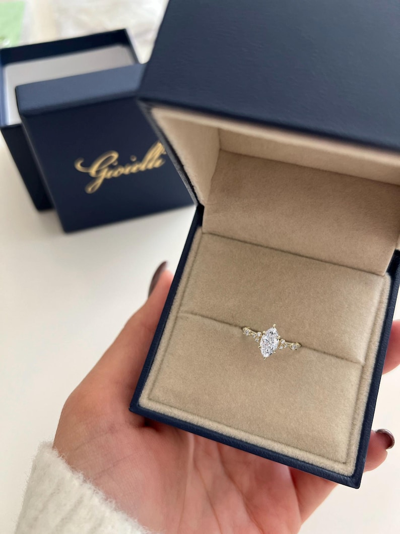 May include: A marquise cut diamond engagement ring with a delicate band and small accent diamonds. The ring is in a blue velvet ring box with the Gioielli logo.