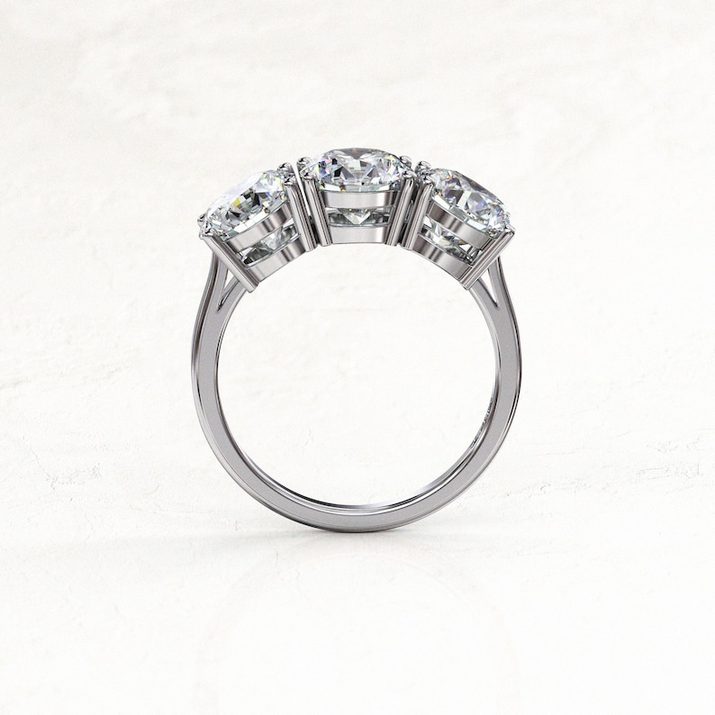 Side profile of a white/silver-colored three-stone moissanite engagement ring, showing the raised round stones, tall prongs, open gallery beneath the settings, and slim polished band.