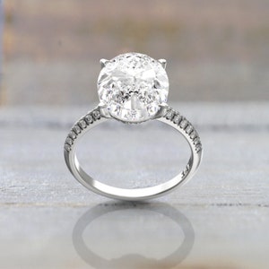 Close-up product image of an oval moissanite engagement ring in white/silver-colored metal, with a prong setting and pavé-set shoulders.