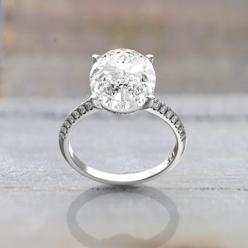 Close-up product image of an oval moissanite engagement ring in white/silver-colored metal, with a prong setting and pavé-set shoulders.