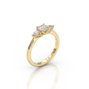 May include: A gold ring with three clear gemstones. The center stone is larger and round, flanked by two smaller, pear-shaped stones. The band is smooth and polished, with a subtle curve. The ring is set against a white background.