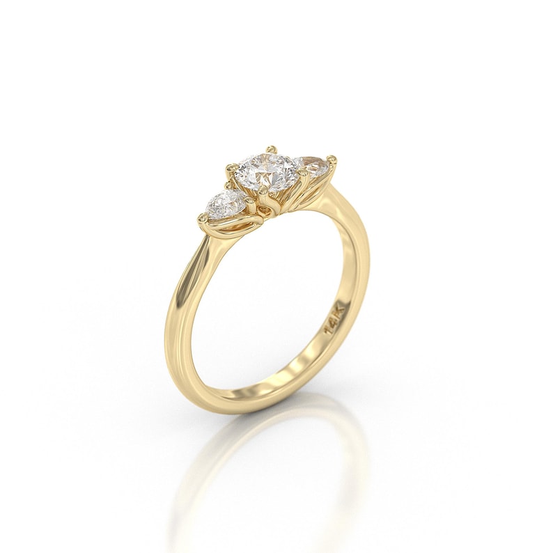 May include: A gold ring with three clear gemstones. The center stone is larger and round, flanked by two smaller, pear-shaped stones. The band is smooth and polished, with a subtle curve. The ring is set against a white background.