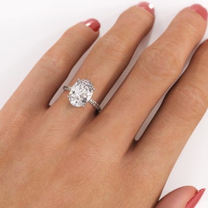 Oval moissanite engagement ring worn on a hand against a white background, with a white/silver-colored metal band and pavé-set shoulders.