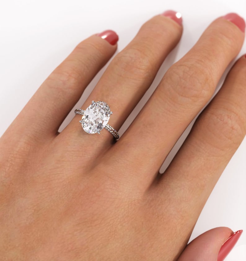 Oval moissanite engagement ring worn on a hand against a white background, with a white/silver-colored metal band and pavé-set shoulders.