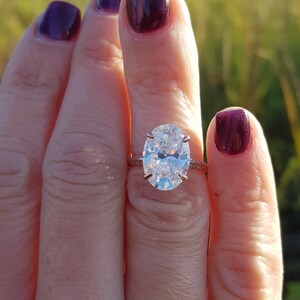 Close-up of an oval moissanite engagement ring held between fingers, showing a prong-set center stone and pavé shoulders.