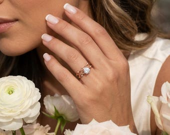 Può includere: Un anello in oro rosa con una pietra preziosa bianca centrale a taglio brillante rotondo incastonata a griffe, affiancata da due pietre bianche rotonde più piccole su ciascun lato, con un delicato design a fascia in metallo intrecciato.