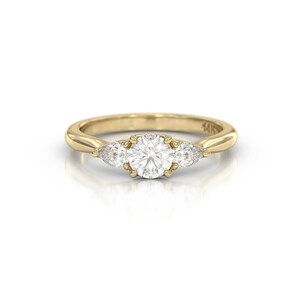 May include: A gold ring with a large, round, clear center stone and two smaller, pear-shaped stones on either side. The ring band is smooth and polished. The ring is set against a white background.