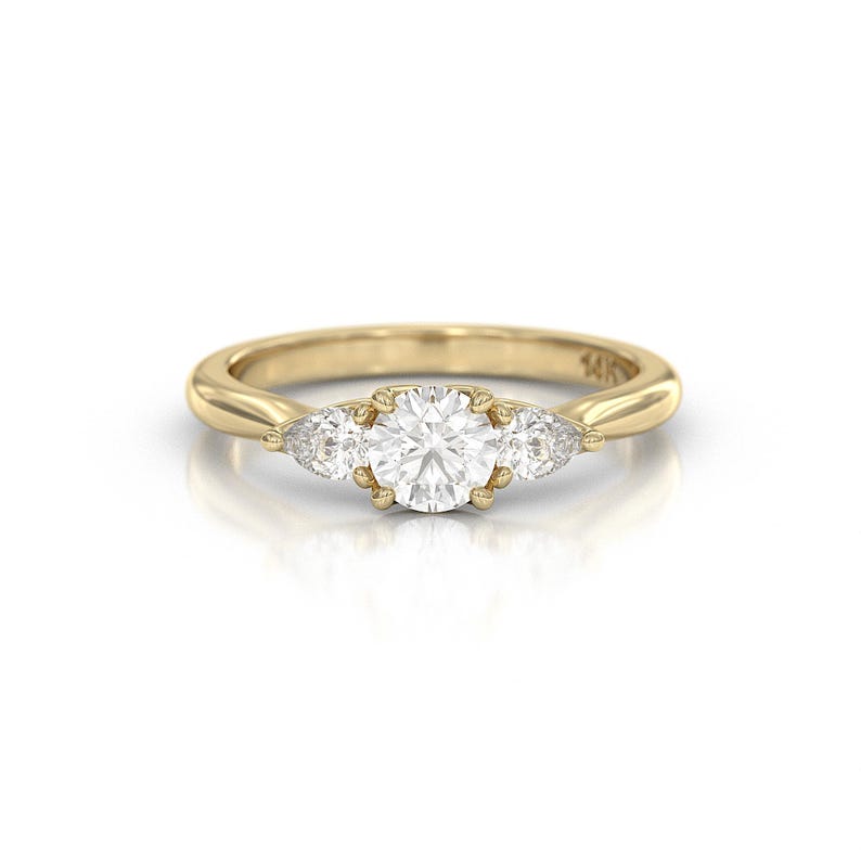 May include: A gold ring with a large, round, clear center stone and two smaller, pear-shaped stones on either side. The ring band is smooth and polished. The ring is set against a white background.