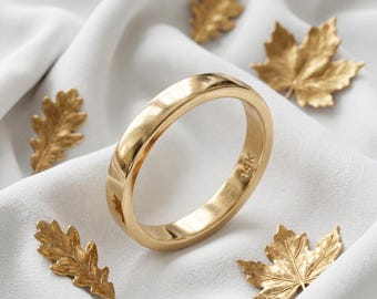 May include: A gold ring with a smooth, rounded design, marked with "14K". The ring is set against a white fabric background, surrounded by gold-colored leaf decorations. The ring is a simple band, perfect for everyday wear.