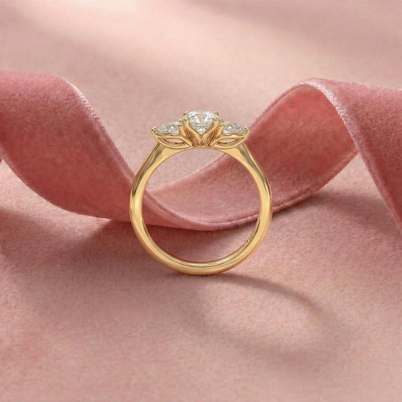 May include: A gold ring with three clear gemstones. The center stone is larger than the two side stones. The ring is set against a pink background with a pink ribbon.