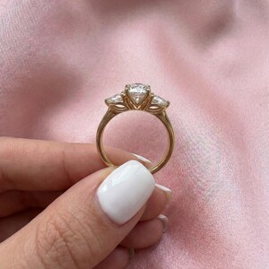 May include: A gold ring with a large, round center stone and two smaller side stones. The ring is held against a pink background, showcasing the ring's design and sparkle. The ring's band is a simple, elegant design.
