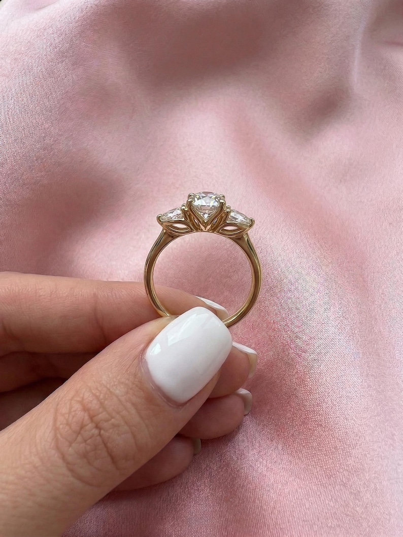 May include: A gold ring with a large, round center stone and two smaller side stones. The ring is held against a pink background, showcasing the ring's design and sparkle. The ring's band is a simple, elegant design.