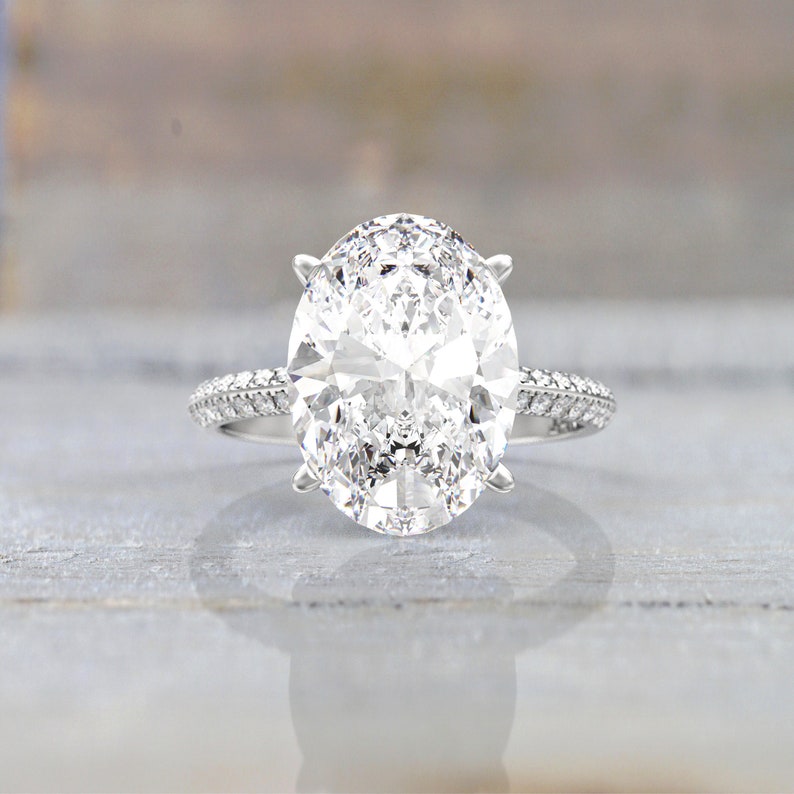 Front view of a white/silver-colored metal engagement ring with a large oval moissanite held by prongs and a pavé-set band visible behind it.