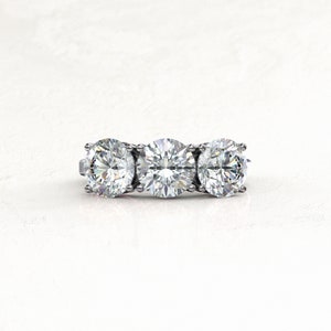 Straight-on view of a white/silver-colored three-stone moissanite engagement ring, with a larger round center stone flanked by two round side stones and secured by rounded prongs.