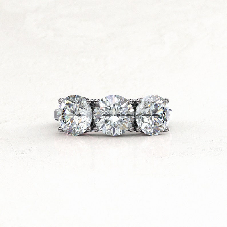 Straight-on view of a white/silver-colored three-stone moissanite engagement ring, with a larger round center stone flanked by two round side stones and secured by rounded prongs.
