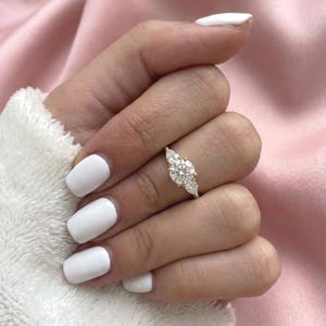 May include: A gold ring with a large, round center stone and two smaller pear-shaped side stones. The ring is set on a hand with white nail polish, against a soft pink background.