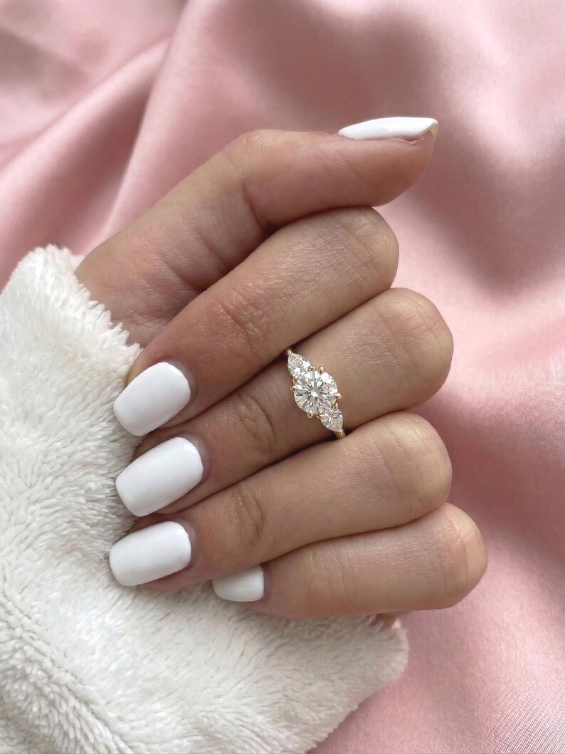 May include: A gold ring with a large, round center stone and two smaller pear-shaped side stones. The ring is set on a hand with white nail polish, against a soft pink background.
