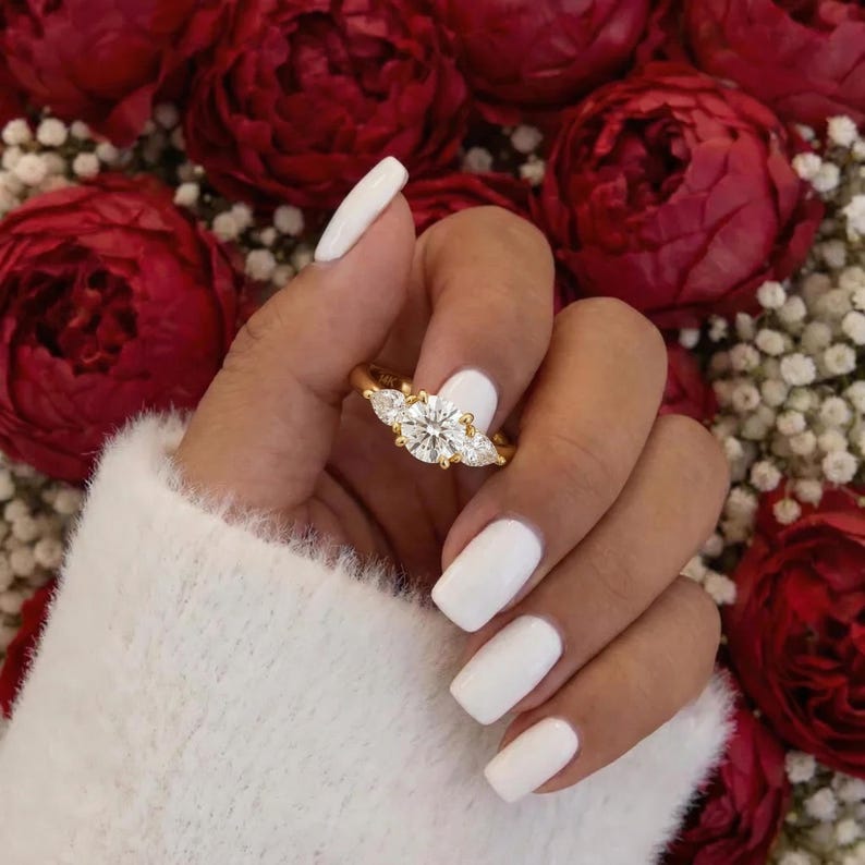 May include: A gold ring with three clear gemstones, a large round center stone flanked by two smaller pear-shaped stones. The ring is held against a backdrop of red flowers and white baby's breath.