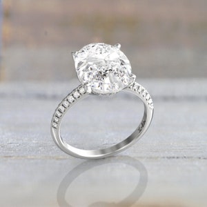 Angled macro view of a white/silver-colored metal engagement ring showing the oval moissanite, prong setting, and pavé stones along both shoulders.