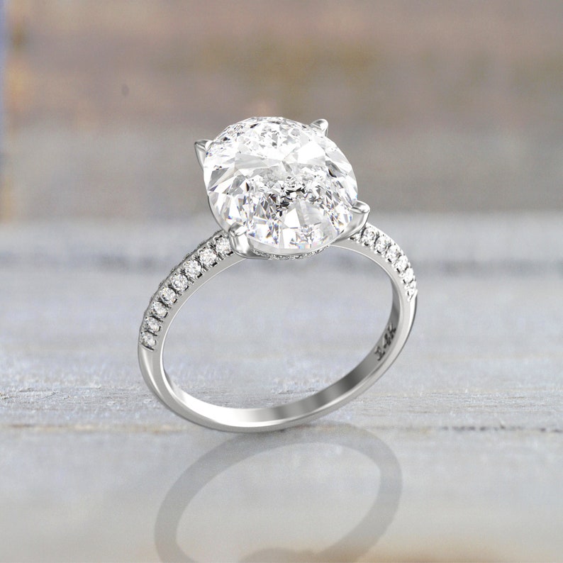 Angled macro view of a white/silver-colored metal engagement ring showing the oval moissanite, prong setting, and pavé stones along both shoulders.