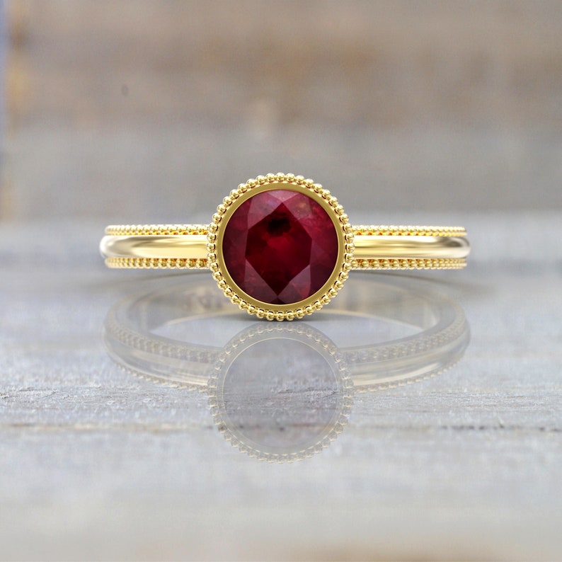 Front view of a yellow gold engagement ring with a large faceted round ruby in a smooth bezel surrounded by a beaded border and a beaded-edge band.