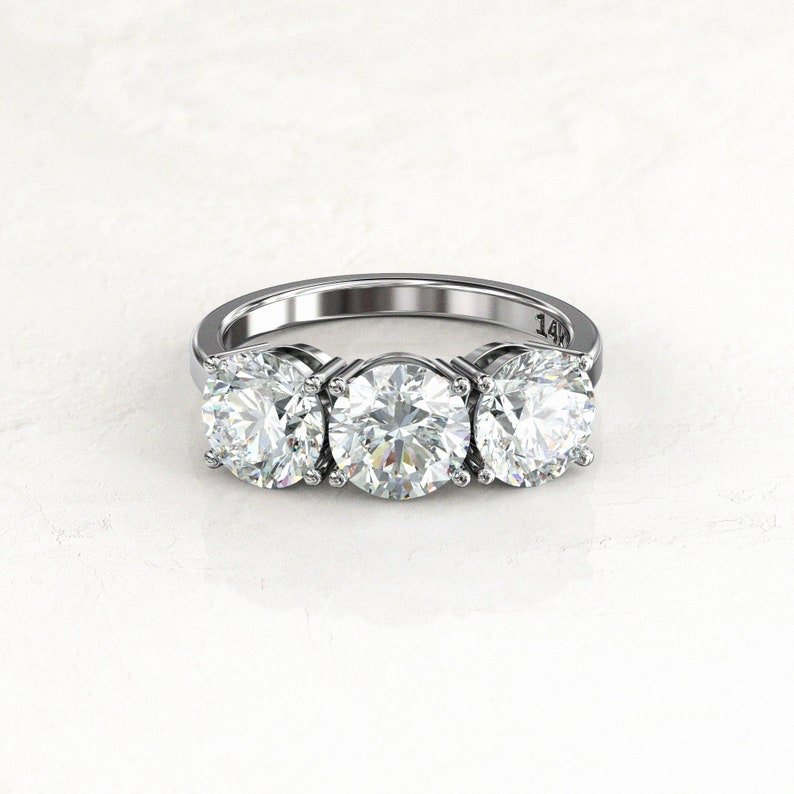 Close front view of three round moissanites in prong settings on a white/silver-colored engagement ring; the inner band is visible behind the stones with a legible 14K stamp.