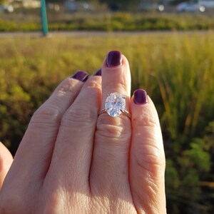 Oval moissanite engagement ring worn on a hand outdoors, showing the elongated center stone, prong setting, and pavé shoulders.