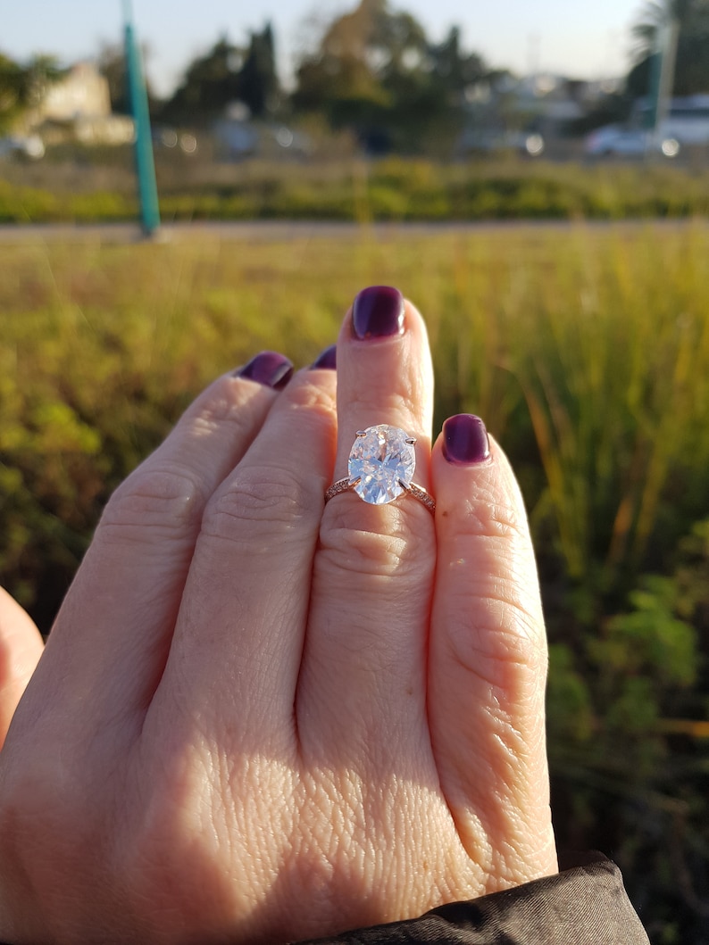 Oval moissanite engagement ring worn on a hand outdoors, showing the elongated center stone, prong setting, and pavé shoulders.