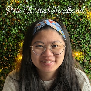 May include: A light blue Pixie Twisted Headband with a cartoon character print. The headband is worn by a person with long, dark hair and round glasses. The background is a green wall of foliage.