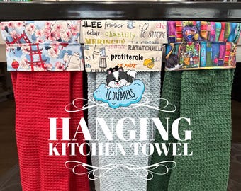 May include: Three hanging kitchen towels with waffle-textured fabric in red, light blue, and green. Each towel features a patterned fabric top with designs including cherry blossoms, French culinary text, and colorful books. Text reads HANGING KITCHEN TOWEL.