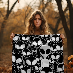 May include: A black and white tapestry featuring a repeating pattern of alien faces. The tapestry is held by a person in front of a blurred background of trees and leaves.