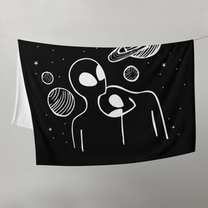 May include: Black and white tapestry featuring two aliens with large eyes and a ringed planet in the background. The tapestry is decorated with white stars on a black background.