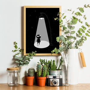 May include: Black and white illustration of a small figure standing in a spotlight against a dark background. The illustration is framed in a wooden frame and displayed on a shelf with plants and a camera.