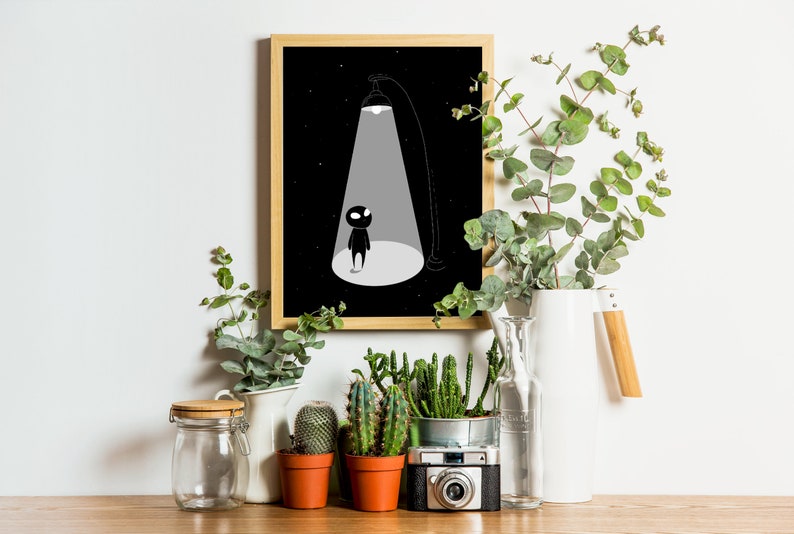 May include: Black and white illustration of a small figure standing in a spotlight against a dark background. The illustration is framed in a wooden frame and displayed on a shelf with plants and a camera.