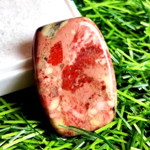 Op de afbeelding: Een gepolijste, rechthoekige edelsteen met een mix van rode, roze en crèmekleurige tinten. De steen heeft een gevlekt uiterlijk met gebieden van effen rood en lichtere roze tinten. Geplaatst op groen gras.