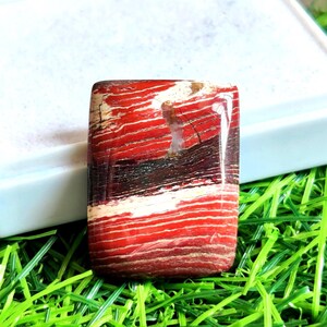 Op de afbeelding: Een rechthoekige edelsteen met een opvallend patroon van rode, witte en donkerbruine horizontale strepen. De gepolijste steen is geplaatst tegen een witte achtergrond en groen gras, waarbij de natuurlijke kleuren en texturen worden getoond.