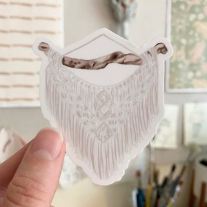 May include: A sticker featuring a macrame wall hanging design. The sticker has a white background with a light brown macrame pattern and a brown wooden dowel. The design includes diamond shapes and fringe.