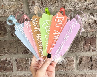 May include: A collection of colorful acrylic bookmarks with inspirational phrases in white script. The bookmarks are in shades of blue, pink, brown, green, orange, and purple, each with a ribbon for hanging. The phrases include "Learn", "Be Still", "Move Mountains", "Revive", "Salt and Light", and "Strong and Courageous".