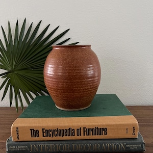 Może przedstawiać: Okrągły, nakrapiany, czerwonobrązowy ceramiczny wazon z szerokim otworem. Wazon stoi na stosie książek z tytułami "The Encyclopedia of Furniture" i "Interior Decoration". Zielony liść palmy znajduje się w tle.