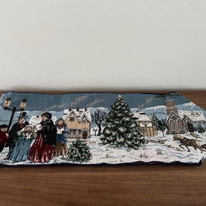 May include: A rectangular Christmas-themed table runner with a winter village scene. The design features figures in period clothing, houses, a church, and a horse-drawn carriage, all set against a snowy backdrop. The runner has blue tassels.