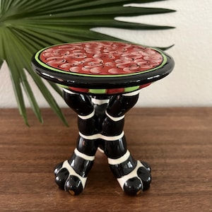 Könnte beinhalten: Ein skurriler Keramik-Sockeltisch mit einer runden Platte mit einem roten Blumendesign. Der Tisch hat schwarze Beine mit weißen Akzenten und pfotenförmigen Füßen. Der Rand ist schwarz mit grünen und roten Streifen.