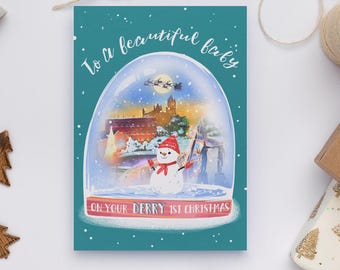 May include: A teal Christmas card with a snow globe design. Inside the globe is a snowman, buildings, and a cityscape. The card reads "To a beautiful baby" and "On your Derry 1st Christmas."