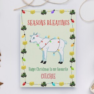 Könnte beinhalten: Eine Weihnachtskarte mit einer weißen Ziege, die eine Lichterkette trägt. Die Karte hat einen grünen Hintergrund mit einem Rand aus Traktoren und Heuballen. Der Text auf der Karte lautet "Seasons Bleatings" und "Frohe Weihnachten an meinen Lieblings-Culchie".