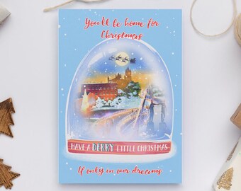 May include: A Christmas card with a blue background features a snow globe design. Inside the globe is a winter scene with buildings, a bridge, and Santa's sleigh. The card reads "You'll be home for Christmas" and "Have a Derry Little Christmas."