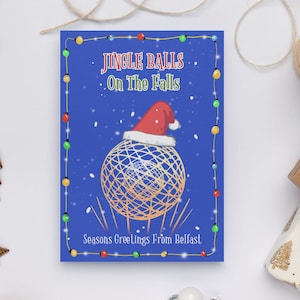 Könnte beinhalten: Eine blaue Weihnachtskarte mit einer weißen und goldenen Kugel, die einen roten Weihnachtsmann-Hut trägt. Die Karte hat einen weißen Rand mit roten, grünen und gelben Lichtern. Der Text auf der Karte lautet "Jingle Balls On The Falls Seasons Greetings From Belfast".