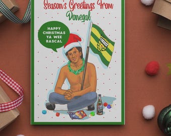 May include: A Christmas card with a green border features a cartoon illustration of a man wearing a Santa hat and holding a flag. The card reads "Season's Greetings From Donegal" and "Happy Christmas Ya Wee Rascal."