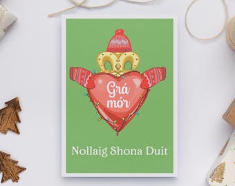 May include: A Christmas card with a green background features a red Claddagh heart design wearing a hat and mittens. The heart has the words "Grá mór" and "Nollaig Shona Duit" in white text. The card is on a white surface.