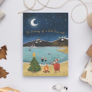 Könnte beinhalten: Eine Weihnachtskarte mit einer skurrilen Illustration einer nächtlichen Strandszene. Die Karte zeigt einen Weihnachtsbaum, ein Lagerfeuer und Menschen, die im Meer schwimmen. Der Text auf der Karte lautet: "I'm Dreaming of a Wild Swim-mas!"